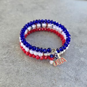 May include: A patriotic bracelet featuring red, white, and blue beads, with a charm that reads "USA". The bracelet is made of three strands of beads, with a mix of faceted and round beads.
