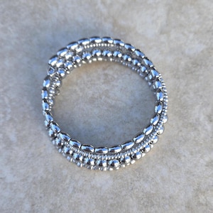 May include: A silver bracelet with three strands of beads. The bracelet is made of small, round beads and larger, oval-shaped beads. The bracelet is circular and appears to be flexible. The jewelry is displayed on a light-colored surface.