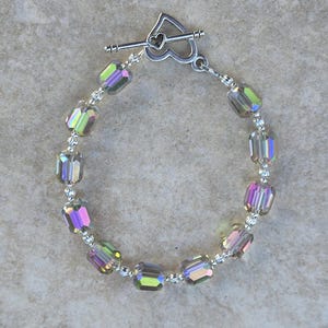 May include: A bracelet featuring iridescent, rectangular beads strung together with small silver-colored beads. The bracelet has a heart-shaped clasp. The beads reflect a range of colors, including purple, green, and yellow.