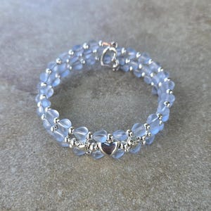 Pode incluir: Uma pulseira delicada com contas azuis claras em forma de coração, unidas por detalhes prateados. A pulseira tem um fecho em forma de coração e um design de duas voltas, criando um efeito em camadas. A joia é exibida em uma superfície neutra.
