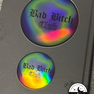 Peut inclure: Deux autocollants holographiques avec le texte "Bad Bitch Club" en police gothique. Les autocollants ont un effet irisé arc-en-ciel. Un petit autocollant est en dessous du plus grand. Les autocollants sont sur une surface gris foncé.
