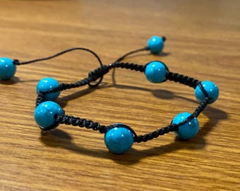 Pode incluir: Um bracelete de contas turquesa com um cordão preto. O bracelete tem sete contas turquesa e um fecho de nó deslizante.