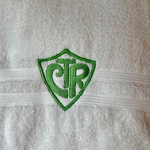 May include: White terry cloth towel with a green embroidered shield design. The shield contains the letters CTR in green. The towel has a textured surface and a decorative stripe.