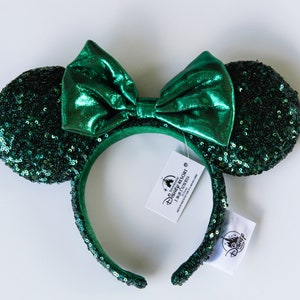 May include: Green sequined mouse ear headband with a matching bow. The ears and headband are covered in small, shiny sequins. A large, shiny green bow sits atop the headband. Disney Resort tags are attached.