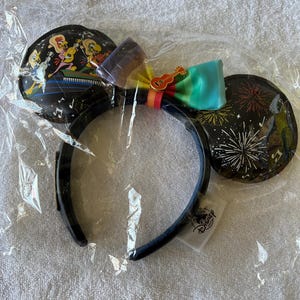 May include: Black Mickey Mouse ear headband with colorful bow and ear details. One ear features a musical scene, the other has fireworks. The bow is rainbow-colored with a guitar charm. The headband is in a clear plastic bag.