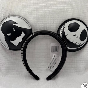 May include: Black and white mouse ear headband featuring characters from The Nightmare Before Christmas. One ear displays a ghost, the other Jack Skellington's face. The headband is black with a striped design.