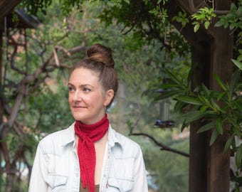 May include: A bright red knitted neckerchief tied in a simple knot around the neck, worn over a dark olive green top and a light blue denim button-down shirt with two chest pockets and a small red tag on the left pocket seam.