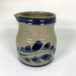 May include: A small, gray ceramic pitcher with a blue rim and a blue floral design. The pitcher has a speckled glaze.