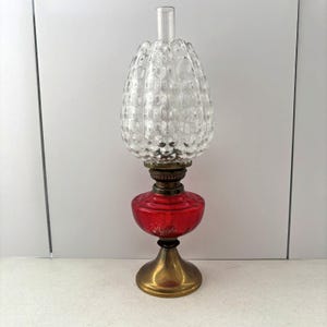 May include: A vintage oil lamp featuring a clear, textured glass shade and a red glass reservoir. It has a brass base and a clear glass chimney. The shade has a distinctive patterned design. This antique lamp is a decorative item.