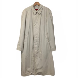 Puede incluir: Una gabardina larga de color beige con cierre de botones y un bolsillo en el pecho. La gabardina tiene un diseño clásico y está hecha de una tela ligera.