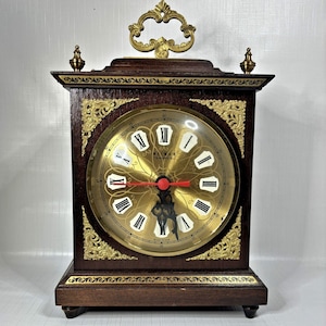 May include: A vintage wooden mantel clock with a gold-tone clock face and Roman numerals. The clock has ornate gold accents, a decorative handle, and two finials. The clock face features the brand name "Wimar".