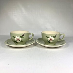 Puede incluir: Dos tazas de té y platillos verdes a juego. Cada taza y platillo está decorado con una flor blanca con detalles en rosa y hojas verdes. Las tazas tienen un asa curva y se asientan sobre un platillo a juego.
