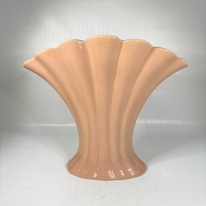 Puede incluir: Un jarrón de cerámica de color rosa pálido con una abertura en forma de abanico y un diseño acanalado. El jarrón tiene un acabado liso y brillante.