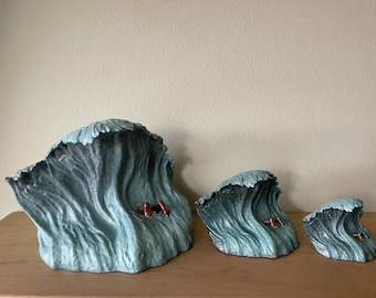 May include: Three ceramic wave sculptures in different sizes, each with a copper-colored surfer figurine inside. The waves are a light blue-green color and have a textured surface.
