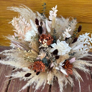 May include: A dried flower bouquet with a mix of white, beige, brown, and rust colored flowers. The bouquet is made up of dried pampas grass, dried roses, and other dried flowers.