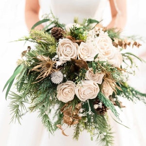 May include: A bridal bouquet featuring cream-colored roses, pine needles, and pine cones. The arrangement includes accents of gold-colored leaves and small white flowers, creating a natural and elegant aesthetic. The bouquet is held against a white background.