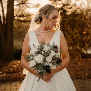 May include: A bride in a white wedding dress holds a bouquet of white roses and pine cones. The bouquet is decorated with sprigs of greenery and berries.