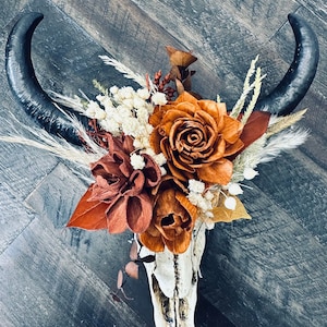 May include: A white animal skull with black horns is decorated with a bouquet of dried flowers. The flowers are in shades of orange, brown, and white.