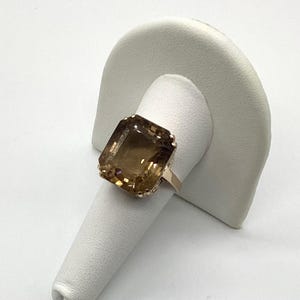 Op de afbeelding: Een gouden ring met een grote, rechthoekige rookkwarts edelsteen. De edelsteen heeft een warme bruine kleur en is gezet in een gouden band. De ring wordt op een witte standaard gepresenteerd.