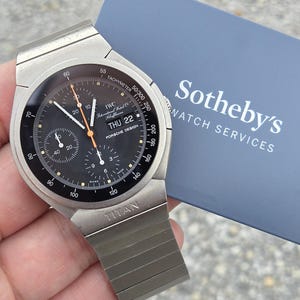 May include: A silver-toned IWC Porsche Design watch with a black face, featuring multiple sub-dials and a tachymeter scale. The watch has a date display and a titanium band. The text "Sotheby's Watch Services" is visible in the background.