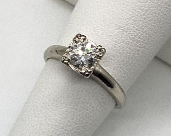 Könnte beinhalten: Ein silberner Ring mit einem großen, klaren, rund geschliffenen Edelstein. Der Edelstein ist in einer quadratischen Fassung mit dekorativen Details gefasst. Der Ring wird auf einer weißen Oberfläche gezeigt.