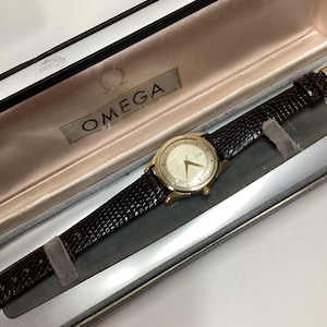 Könnte beinhalten: Eine goldene Armbanduhr mit weißem Zifferblatt und schwarzem Lederarmband in einer braunen Samtbox mit dem Wort "OMEGA" auf der Innenseite.