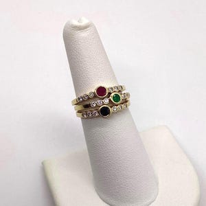 Könnte beinhalten: Ein Stapel aus drei Goldringen, von denen jeder einen runden Edelstein in Rot, Grün beziehungsweise Dunkelblau in der Mitte trägt. Jeder Ring ist mit einer Reihe kleiner, klarer, rund geschliffener Steine verziert, die entlang der Ringschiene gefasst sind.
