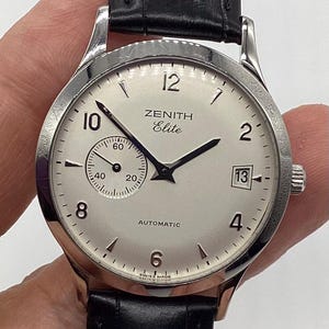 May include: A silver Zenith Elite automatic wristwatch with a white face, black hands, and a black leather band. The watch face displays the Zenith logo, a small seconds dial, and the date.