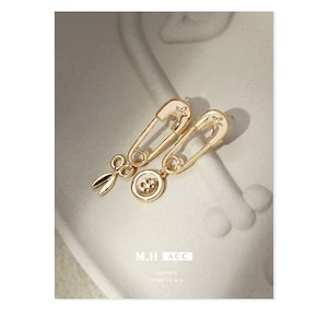 May include: Gold-tone safety pin earrings with a button and a pair of scissors charm. The earrings are on a white background.
