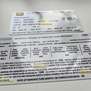 Op de afbeelding: Close-up van een wit label met zwarte tekst met details over de motorspecificaties voor een zware dieselmotor uit modeljaar 1999. Het label bevat informatie over de cilinderinhoud van de motor, de klepspeling, het brandstofverbruik en het maximale vermogen. De tekst "IMPORTANT ENGINE INFORMATION/AVIS IMPORTANT" staat bovenaan het label.
