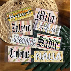 May include: A collection of rectangular plastic luggage tags featuring various floral and tropical patterns. Each tag displays a unique name in distinct typography, including Briani, Mila, Zalynn, Leilani, Jasmine, Sadie, Taylynne, and Nalia.