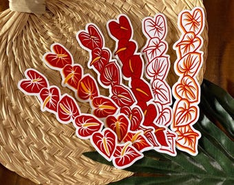 May include: A collection of red and white anthurium flower stickers with yellow accents. The stickers are arranged on a woven straw fan and a green leaf. The stickers have a white border.