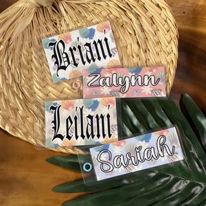 May include: Four rectangular clear plastic name tags featuring colorful seashell and floral patterns. The tags display the names Briani, Zalynn, Leilani, and Sariah in black script or gothic-style fonts, each with a small colored ring at the edge.