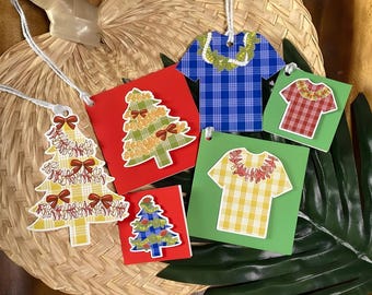 May include: A collection of colorful, decorative gift tags featuring Christmas trees and Hawaiian shirts. The tags are made of paper and have string attached for hanging. The background includes a woven fan and tropical leaves.