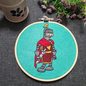 May include: Embroidered hoop art featuring a robot in a red and gold knight costume holding a sword. The robot has a silver helmet and a blue face. The background is turquoise.