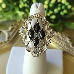 May include: An ornate, gold-toned ring with a central design of four black, oval-shaped stones. The ring is surrounded by clear, round crystals and sits on a white ring display. The ring is displayed on a gold-colored, decorative tray.