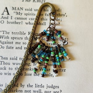 May include: A vintage-style bookmark with a decorative beaded tassel. The bookmark is made of a bronze-colored metal with an ornate design. The tassel features a mix of colorful beads in shades of blue, green, brown, and white. The bookmark is resting on a page of an old book.
