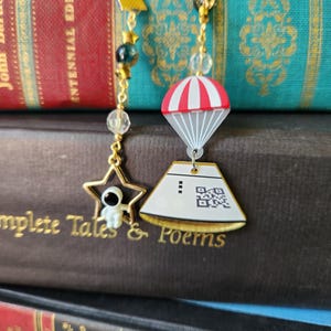 Puede incluir: Dos marcadores dorados con colgantes. Uno presenta una estrella con un astronauta, el otro un diseño de paracaídas. Ambos tienen cuentas transparentes y azules. Los marcadores están sobre una pila de libros con letras doradas.