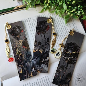 Puede incluir: Cuatro marcapáginas decorativos con diseños de rosas negras y detalles dorados. Cada marcapáginas presenta una ilustración de rosa diferente, con cadenas y abalorios dorados. Los marcapáginas se muestran en un libro abierto con texto.