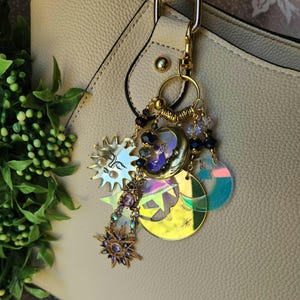 Pode incluir: Um berloque de bolsa com tema celestial com detalhes em metal dourado. O berloque apresenta um design de sol, lua e estrela com detalhes iridescentes e em tons de joia. O berloque está preso a uma bolsa bege.
