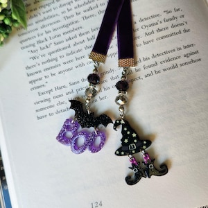 May include: Two Halloween-themed bookmarks with purple velvet ribbons. One features a black glitter bat and the word "BOO" in purple glitter. The other has a witch hat and legs with black boots.