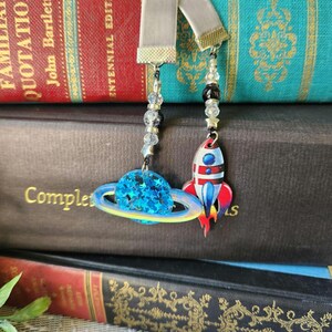 Puede incluir: Dos marcapáginas con temática espacial con cintas de terciopelo gris. Uno presenta un planeta azul con anillos amarillos, el otro un cohete rojo y blanco. Ambos tienen cadenas de cuentas y dijes de estrella, sobre una pila de libros antiguos.