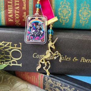 May include: A bookmark with a pink velvet ribbon and a gold-colored clasp. The bookmark features a clear acrylic charm with a skeleton riding a unicorn under a rainbow, with the text "THE AUDACITY." A gold-colored skeleton unicorn charm hangs below.