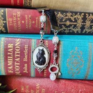 May include: A black velvet ribbon bookmark featuring two silver-toned metal chains. One chain holds an oval pendant with a black cat silhouette on a white background. The second chain features a silver cat-shaped charm, white pearls, and black beads.