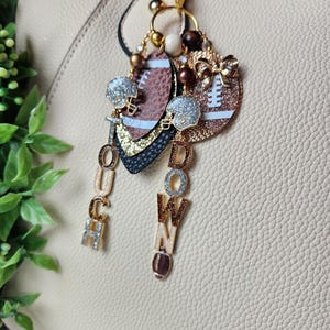 May include: A football-themed keychain with a beige background. The keychain features two football-shaped charms, a glittery football helmet, and the word "TOUCHDOWN" spelled out in individual letters. The charms are adorned with gold and brown accents.