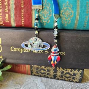 Puede incluir: Dos marcadores de cinta de terciopelo azul con colgantes colgantes. Un colgante es un planeta con purpurina, el otro es un cohete rojo, blanco y azul. Los marcadores cuelgan sobre una pila de libros.