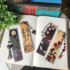 Puede incluir: Cuatro marcapáginas decorativos con diseños únicos, colocados sobre un libro abierto. Uno presenta una casa embrujada bajo la luna llena, otro esqueletos bailando, y dos más muestran patrones florales con rosas. Cada marcapáginas tiene una cadena decorativa.