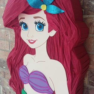 Puede incluir: Una pi&ntilde;ata con forma de sirena de dibujos animados con largo cabello de papel crep&eacute; rojo, un accesorio para el cabello azul brillante, un top de concha morado brillante y una cola verde brillante. La figura tiene grandes ojos azules.