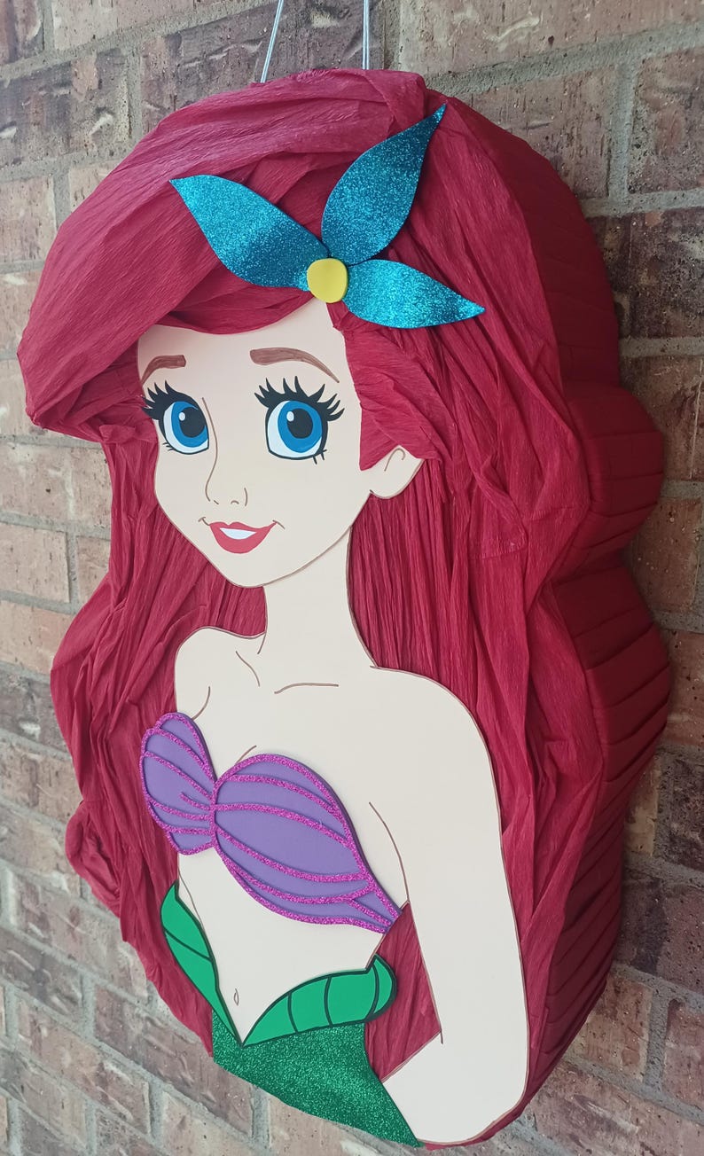 Puede incluir: Una pi&ntilde;ata con forma de sirena de dibujos animados con largo cabello de papel crep&eacute; rojo, un accesorio para el cabello azul brillante, un top de concha morado brillante y una cola verde brillante. La figura tiene grandes ojos azules.
