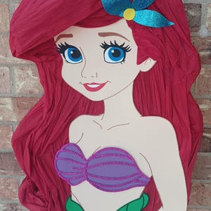 Puede incluir: Una pi&ntilde;ata con forma de sirena de dibujos animados con cabello largo y voluminoso de papel crep&eacute; rojo, un accesorio para el cabello de color verde azulado brillante, un top de concha morado brillante y una cola verde brillante.